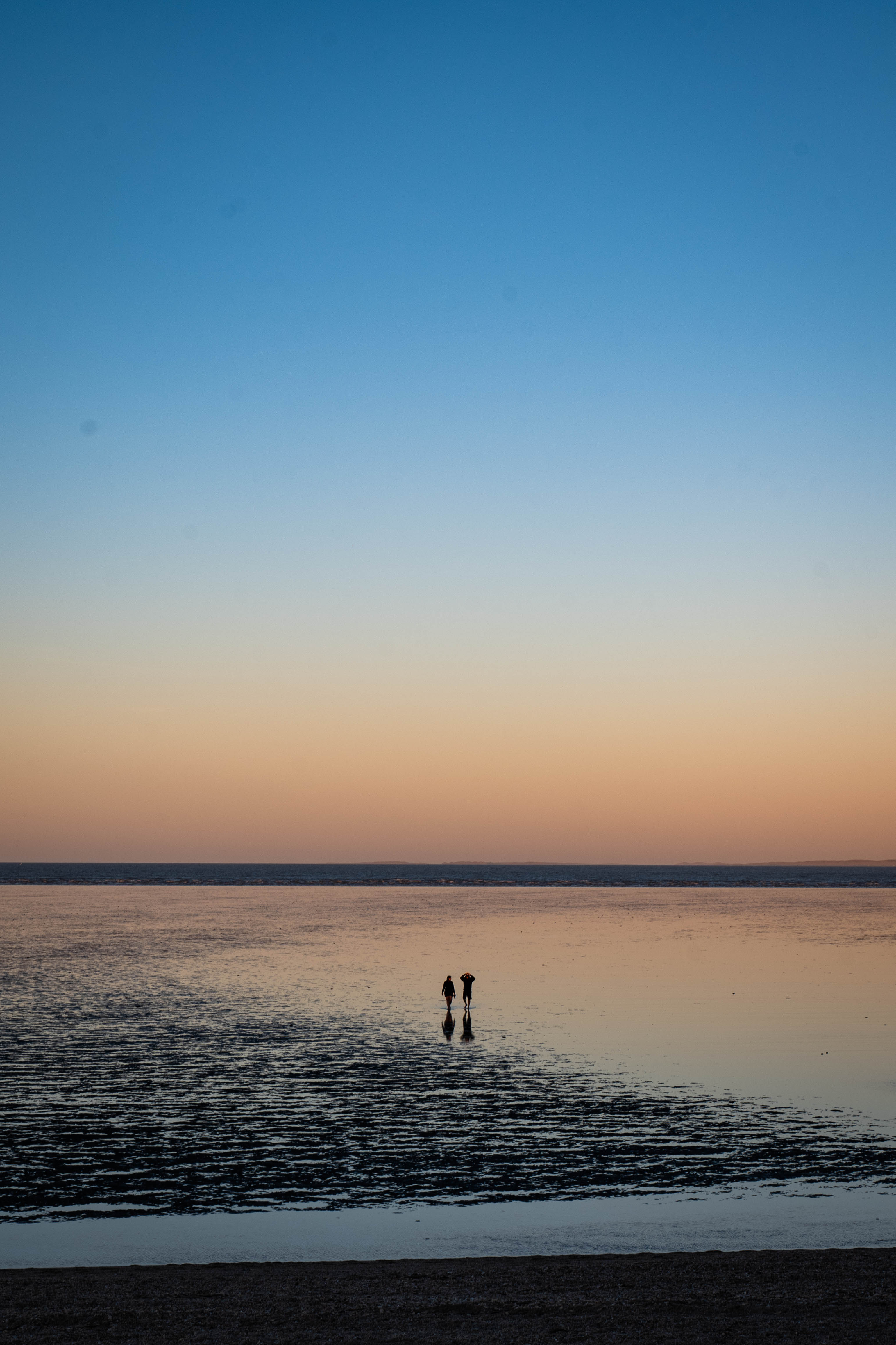 Two people walking across shallow tidal water beneath a pastel evening sky