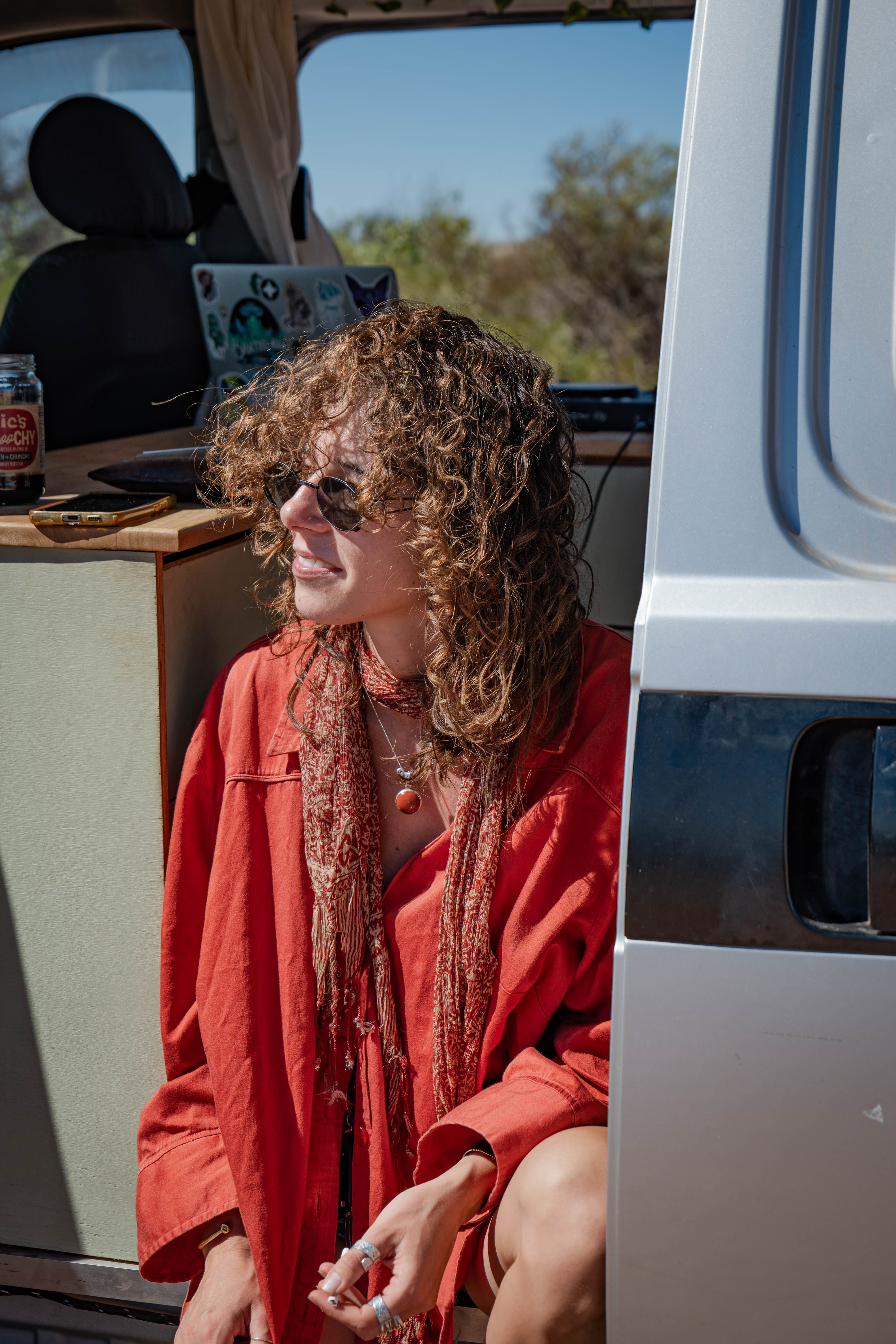 A curly-haired woman in a rust-red shirt sitting in the doorway of a campervan