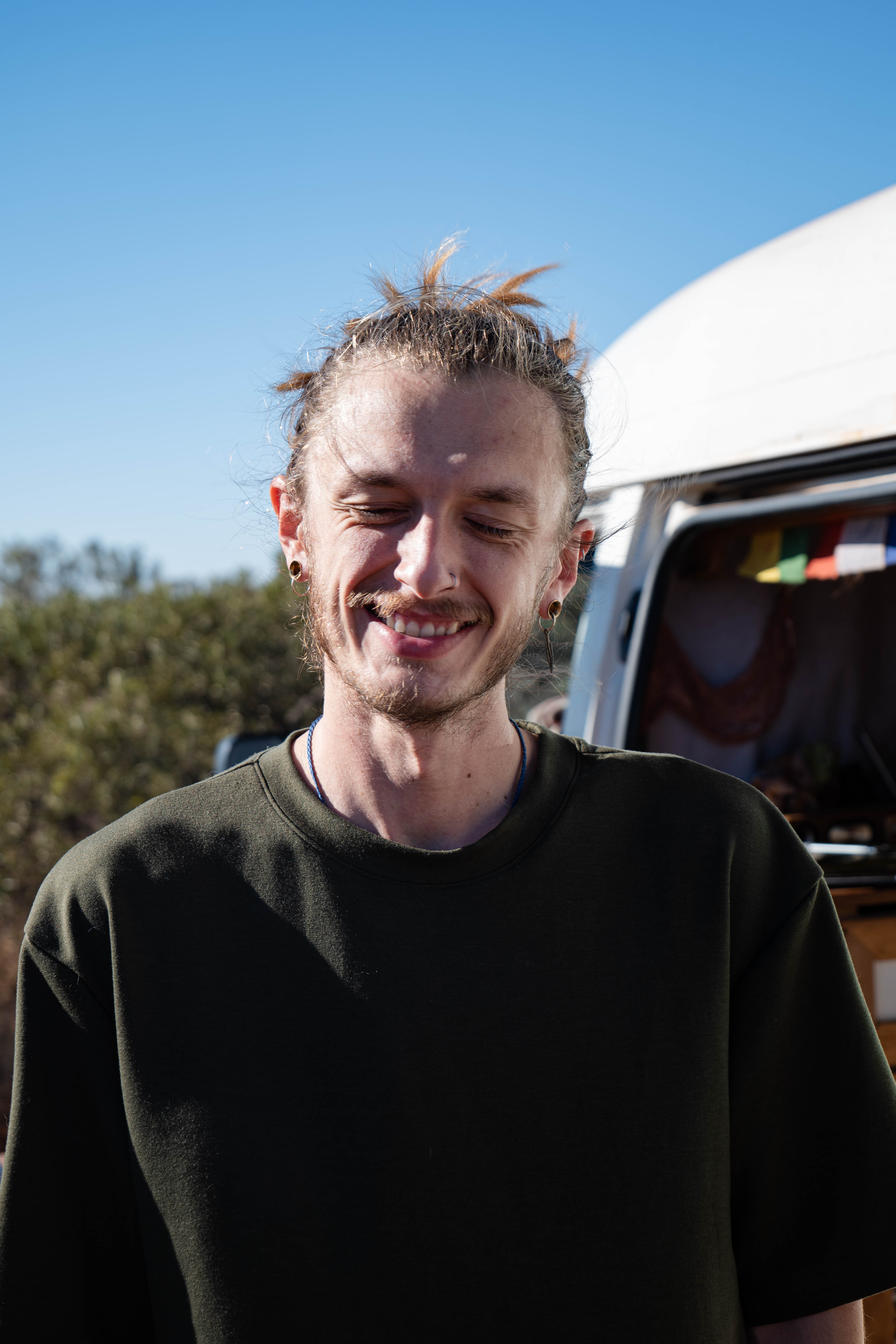 A smiling man with tied-back hair standing beside a campervan